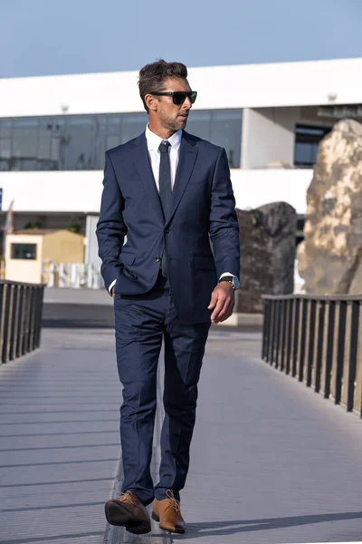 Full body self assured male entrepreneur in suit and sunglasses holding hand in pocket and looking away while walking on footbridge on sunny day in city