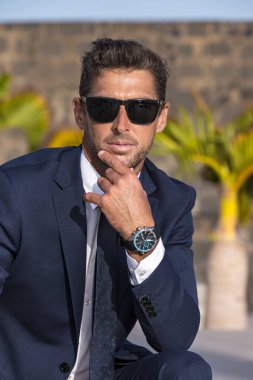 Serious businessman in suit with stylish sunglasses and expensive wristwatch touching chin and looking at camera on blurred background of tropical palms and stone wall on city street