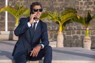 Takım elbiseli ve güneş gözlüklü bir erkek girişimci şehrin sokaklarında tropikal palmiyelerin ve taş duvarların yanında otururken gözlerini kaçırıp akıllı telefondan konuşuyor.