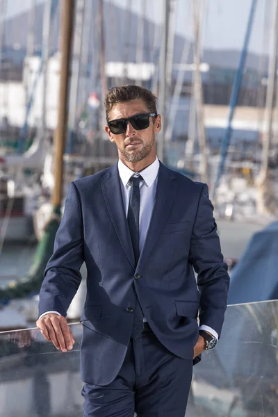 Self assured businessman in suit and sunglasses holding hand in pocket and looking at camera while leaning on fence on blurred background of yachts in city harbor.