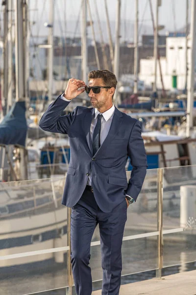 Male entrepreneur with hand in pocket of suit adjusting sunglasses and looking away while standing on embankment against yachts on sunny day in city