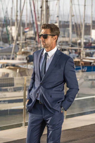 Takım elbiseli ve güneş gözlüklü kendine güvenen işadamı güneşli bir yaz günü şehrin limanında setin kenarında dikilirken el ele tutuşup gözlerini kaçırıyor..