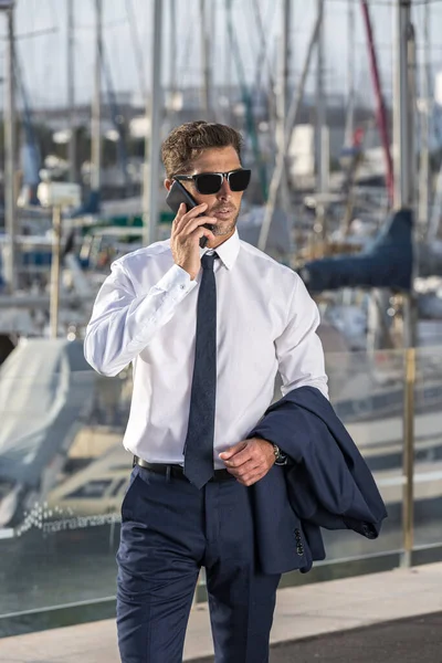 Resmi giyinmiş, güneş gözlüklü, ceketli, deniz kenarında yürürken ve güneşli bir limanda akıllı telefon görüşmeleri yaparken gözlerini kaçıran zeki bir yönetici.