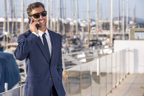 スーツ姿の幸せな実業家とサングラスを笑顔で 街の港で晴れた夏の日を過ごしながらスマートフォンで話しています ロイヤリティフリーのストック画像