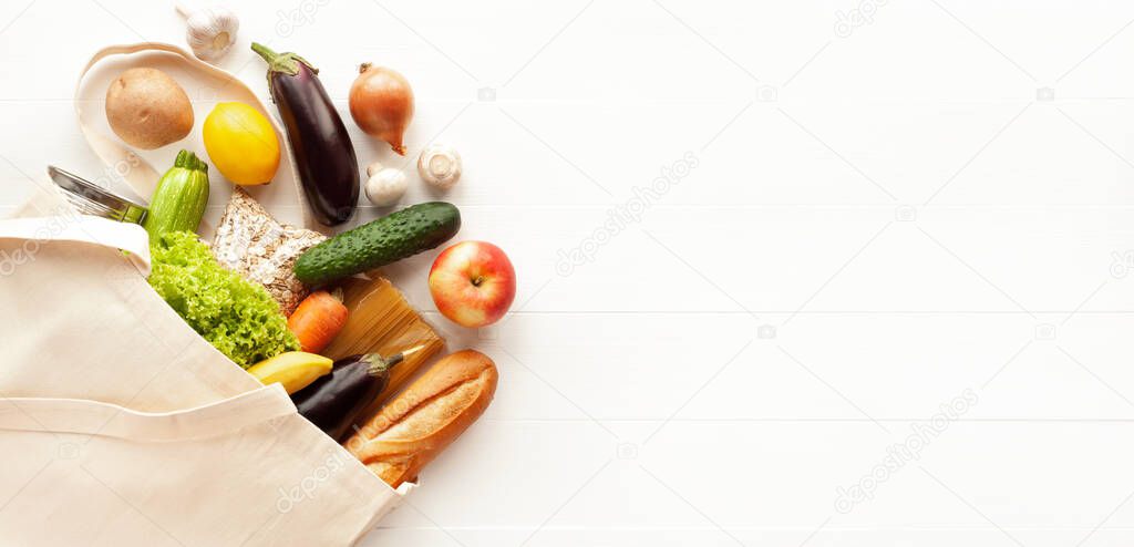 Cotton grocery tote bag with fresh vegetables, fruits, baguette and canned goods on white wooden background. Healthy food shopping, eco-friendly concept. Flat lay, copy space, banner.