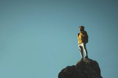 Sarı ceketli, sırt çantalı ve gözlüklü bir adam mavi gökyüzü arka planında bir kayanın tepesinde duruyor..
