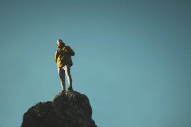 Sarı ceketli ve gözlüklü bir adam mavi gökyüzü arka planında bir kayanın üstünde duruyor..