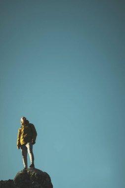 Sarı ceketli ve gözlüklü bir adam mavi gökyüzü arka planında bir kayanın üstünde duruyor..