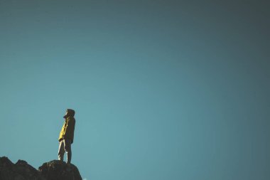 Sarı paltolu ve gözlüklü bir adam bir kayanın üstünde duruyor ve mavi bir arka planda güneşe bakıyor..