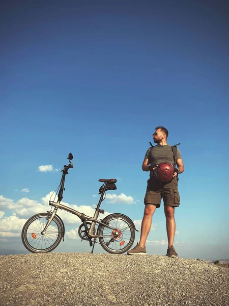 A man with a foldable bicycle shirts, sunglasses and a helmet on a blue sky background.