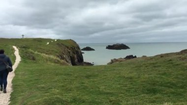 A plus-sized woman walks the trail along the coastal cliffs of Ynys Llanddwyn island. The sea in the background with vibrant green grass and a religious cross in the distance. Anglesey, Wales, UK