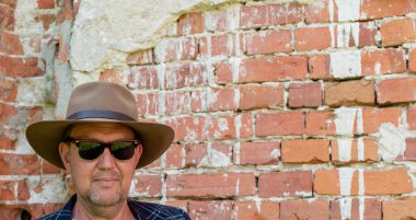 Eski kırmızı tuğla duvarın bulanık arka planında şapka ve güneş gözlüğü takan orta yaşlı bir adamın portresi.