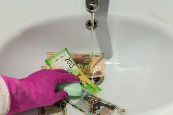 Hand in glove with soap on banknotes of russian rubles close-up under water jet in white ceramic wash basin. View from above