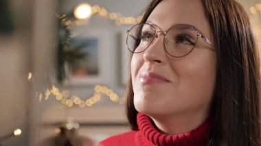 Noel ve Yeni Yıl Arifesi. Mutlu kadın Noel ağacına hayranlıkla bakıyor, Noel çelengi ışıkları gözlüklere yansıyor. Yakın çekim ve yavaş çekim