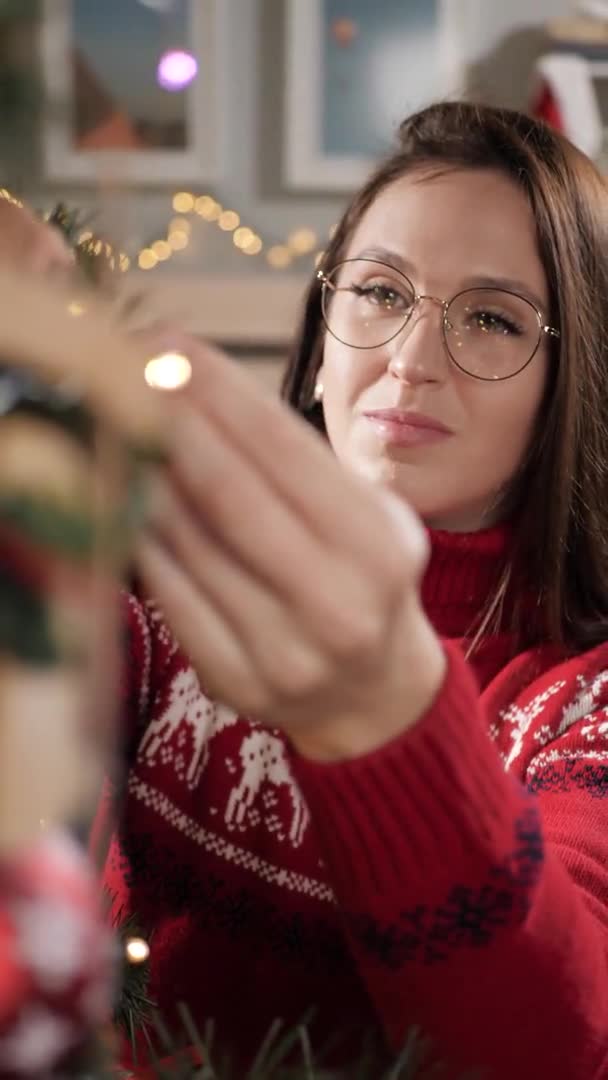 A decorar a árvore de Natal. A visão vertical da mulher feliz atraente em copos e suéter vermelho decora a árvore de Natal, põe o brinquedo de Natal no ramo. Close-up e câmera lenta — Vídeo de Stock