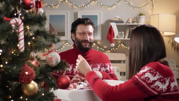 Een stel versiert de kerstboom. Gelukkig glimlachende man en vrouw in rode trui zitten op de vloer praten en zet kerstspeelgoed op takken van de kerstboom. Langzame beweging — Stockvideo