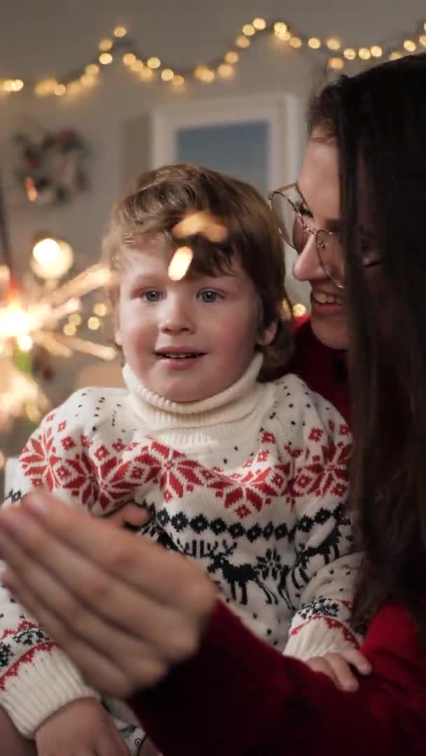 Julelys, stjernekastere. Lodret visning af glad smilende kvinde og barn 2-3 år gammelt kig på stjernekaster, julekrans i baggrunden. Close-up og slow motion – Stock-video