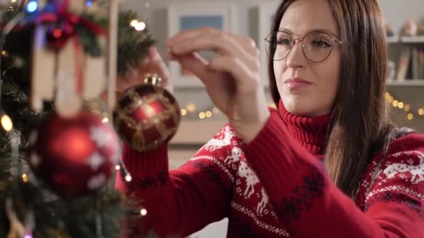 Vrouw is kerstboom aan het versieren. Jonge aantrekkelijke vrouw in bril en rode trui versiert kerstboom, zet ze kerstspeelgoed op tak. Langzame beweging — Stockvideo