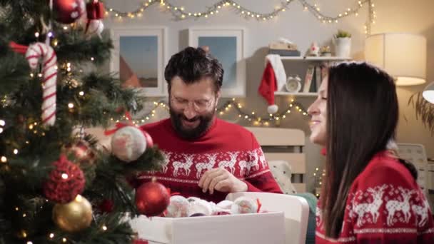Een stel versiert bomen en rotzooit wat. Gelukkig glimlachende man en vrouw in rode trui versieren kerstboom, rotzooien rond lachen en gooien speelgoed naar elkaar. Langzame beweging — Stockvideo