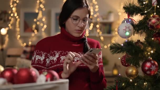Kvinna som jobbar på julafton. Pensiv kvinna dekorerar julgran och distraheras av att skriva meddelande i smartphone — Stockvideo