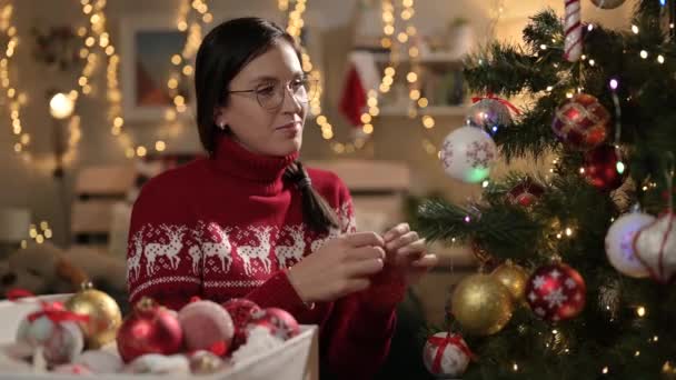 Wanita menghias pohon Natal. Wanita tersenyum dengan sweater merah menghias pohon Natal, mengambil mainan dari kotak dan meletakkannya di cabang, lampu karangan bunga di latar belakang. Konsep malam Natal — Stok Video