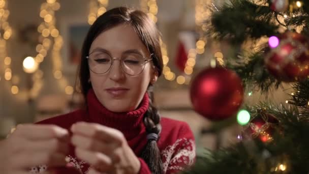 Menghias pohon Natal. Wanita bahagia dengan sweater merah janda Pohon Natal dan menempatkan mainan di cabang pohon Natal pada malam Natal. Close-up — Stok Video