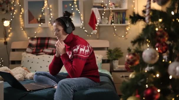 Jouluaattona nainen puhuu videopuhelussa. Huoneessa koristeltu jouluksi puu ja valot seppeleitä nainen istuu sängyllä ja puhuu kannettavan videopuhelu — kuvapankkivideo