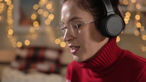 Na mulher de Natal com fones de ouvido está falando em videochamada, luzes de guirlandas estão brilhando no fundo. Close-up — Vídeo de Stock