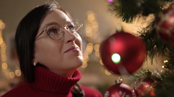 Szenteste. Boldog nő néz karácsonyfa és örül, karácsonyi fények koszorúk tükröződnek szemüveg. Közelkép — Stock videók