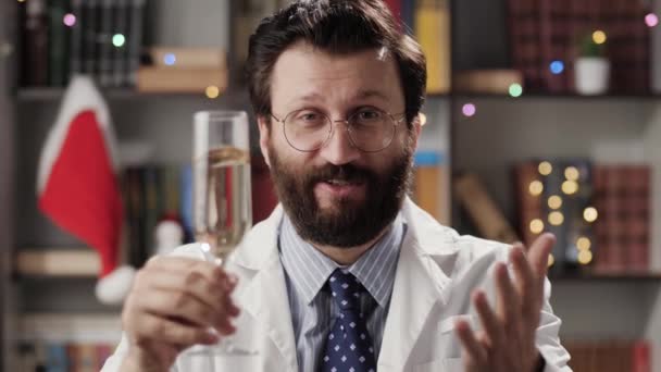 Счастливого сочельника, счастливого Нового года. Мужчина-врач в очках и белом халате с шампанским в руке, глядя в камеру и произнося поздравительную речь. Медленное движение — стоковое видео