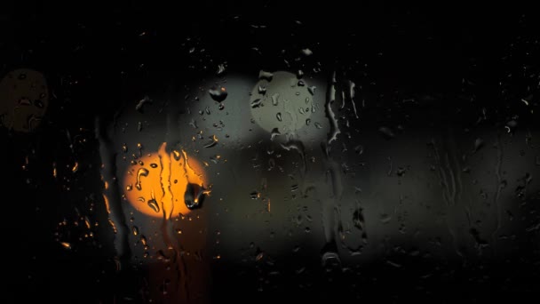Az esőcseppek lassan csepegnek az üvegre, és éjszaka nagy fényfoltok hátterében folynak le. Közelkép — Stock videók