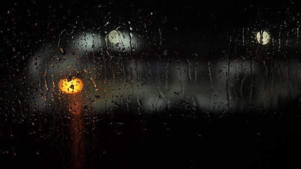 Des gouttes de pluie sur verre. La nuit, sur fond de taches de lumière, gouttes d'eau goutte à goutte sur verre et couler vers le bas, il pleut. Gros plan — Video