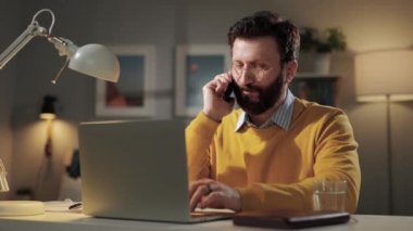 Adam telefonda konuşuyor ve çalışıyor. Dost canlısı gülümseyen sakallı adam telefonla konuşuyor ve bilgisayar ve kağıt üzerinde notlar alıyor.