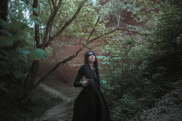 Young woman in witch stands in black dress in dark and foggy forest. halloween costume for dark party.