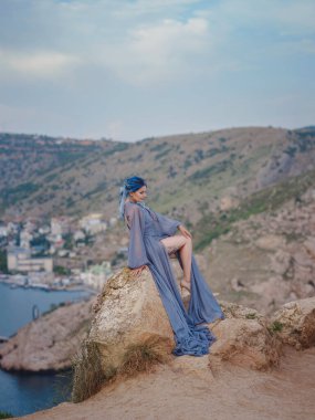 Gri elbiseli, mavi saçlı genç ve güzel bir kadın deniz dağlarına bakıyor. Doğadaki kadın. Moda fotoğrafları. Deniz perisi rüzgarı dinliyor.