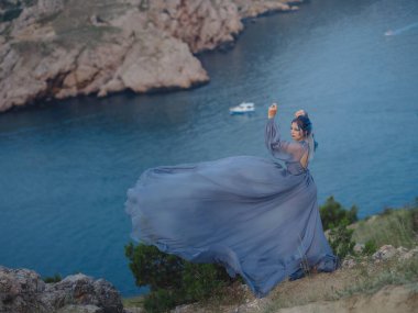 Gri elbiseli, mavi saçlı genç ve güzel bir kadın deniz dağlarına bakıyor. Doğadaki kadın. Moda fotoğrafları. İpek Kumaş Rüzgarda Dalgalanıyor