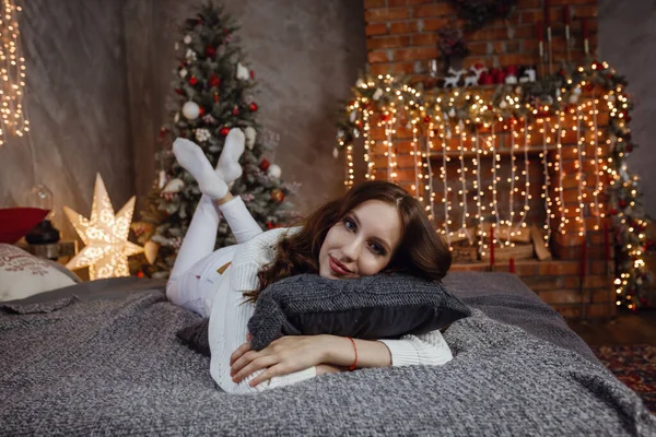 漂亮的模特 身材完美 穿着舒适的毛衣 坐在室内装饰的床上过年 穿着时髦舒适衣服的年轻女人躺在床上 靠着阁楼里的圣诞树 — 图库照片