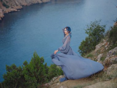 Gri elbiseli, mavi saçlı genç ve güzel bir kadın deniz dağlarına bakıyor. Doğadaki kadın. Moda fotoğrafları. İpek Kumaş Rüzgarda Dalgalanıyor