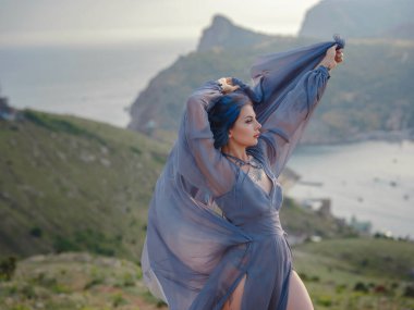 Gri elbiseli, mavi saçlı genç ve güzel bir kadın deniz dağlarına bakıyor. Doğadaki kadın. Moda fotoğrafları. İpek Kumaş Rüzgarda Dalgalanıyor