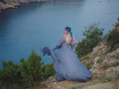 Gri elbiseli, mavi saçlı genç ve güzel bir kadın deniz dağlarına bakıyor. Doğadaki kadın. Moda fotoğrafları. İpek Kumaş Rüzgarda Dalgalanıyor