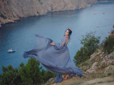 Gri elbiseli, mavi saçlı genç ve güzel bir kadın deniz dağlarına bakıyor. Doğadaki kadın. Moda fotoğrafları. İpek Kumaş Rüzgarda Dalgalanıyor