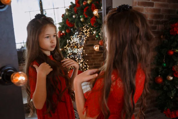 鏡の近くにポーズをとる赤いドレスのかわいい女の子 冬休みの季節 子供の頃 ロフトスタイルのインテリア背景にクリスマスの装飾 — ストック写真