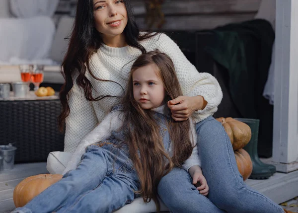 Young mother and daughter sitting on wooden steps of porch decorated with pumpkins. Autumn sunny lifestyle portrait.