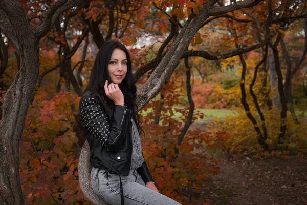 年轻的白人女子穿着黑色皮夹克 灰色毛衣和牛仔裤在秋天的公园里 温暖的天气 — 图库照片
