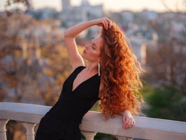 Siyah takım elbiseli kızıl genç kadın bahar şehrinde yürüyor. İş kadını iş gününden sonra gün batımının tadını çıkarıyor.