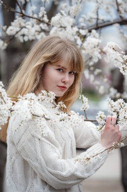 Bahar zamanı kiraz çiçeği bahçesinde beyaz süveter giyen güzel genç bir kadın. Açık hava moda fotoğrafı.