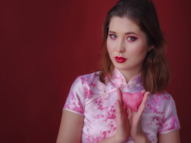 Çekici Asyalı kadının portresi. Elinde küçük parlak bir kalp tutuyordu. Kırmızı arka planda, geleneksel Çin elbisesi giymişti.