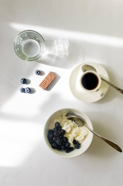 Healthy breakfast bowl with cottage cheese, yogurt, blueberries and cup of hot coffee in the morning sunlight with window shadow on white table.