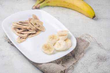 Olgun çiğ muz ve kurutulmuş muz dilimleri açık gri arka planda tabakta cipsler. Meyve cipsi. Sağlıklı beslenme konsepti, atıştırmalık, şekersiz. Üst görünüm, alanı kopyala.