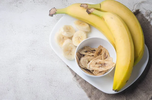 Ripe raw banana and dried banana slices chips in plate on light grey background. Fruit chips. Healthy eating concept, snack, no sugar. Top view, copy space.